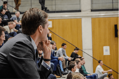 A man in a suit sits in an auditorium, watching a presentation with others seated around him.