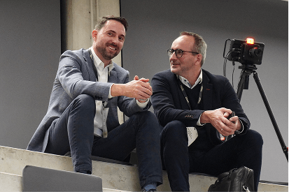 Two men sit on an upper row of steps, chatting near a video camera setup.