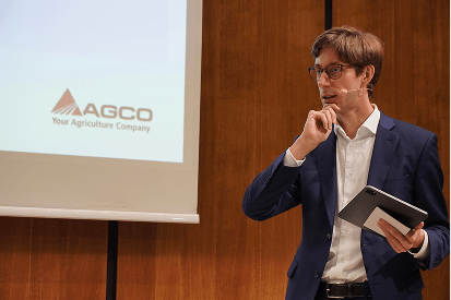 A presenter in a suit speaks on stage beside a screen.