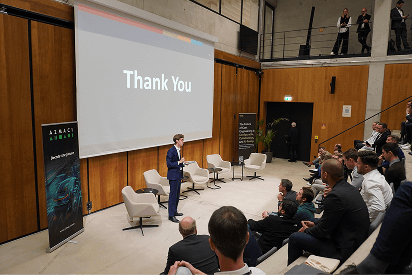 A speaker addresses an audience in a modern auditorium, with a large screen behind him showing the words “Thank You.”
