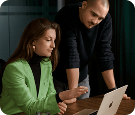 A man and woman in business casual attire collaborating at a wooden table, looking at a MacBook with a focused expression.