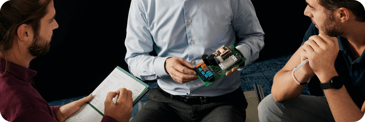 Three men in discussion, one holding a circuit board while the others take notes and ask questions.
