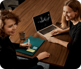 Two women smiling and collaborating at a wooden desk with a laptop, notebook, and glass of water.