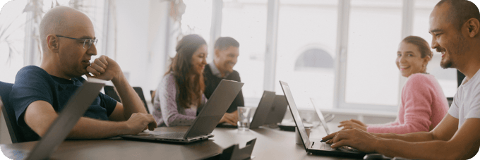 Team members working together at a meeting table, smiling and engaged with laptops