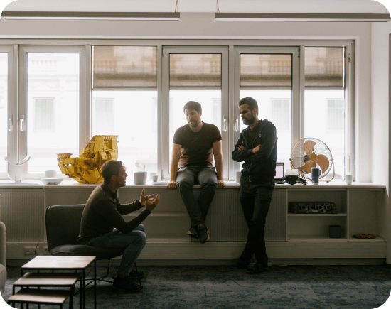 Three team members having a casual discussion by the window in a relaxed office setting.