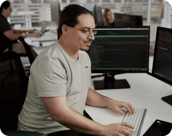 Software engineer typing at a desk with dual monitors displaying code in a collaborative workspace.
