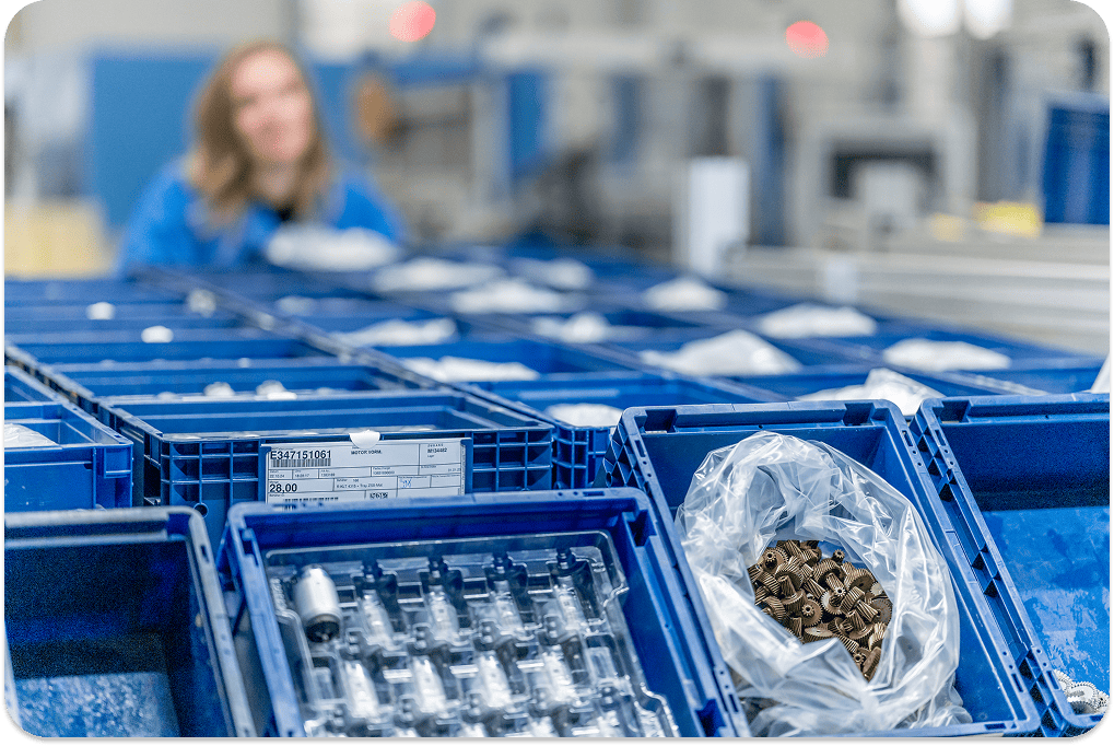 Close-up of labeled blue crates filled with industrial components in a production warehouse, with a blurred worker in the background Close-up of labeled blue crates filled with industrial components in a production warehouse, with a blurred worker in the background
