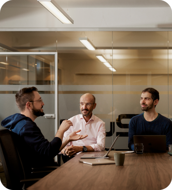 Three colleagues engaged in a discussion around a meeting table with laptops and coffee cups in a modern office setting.