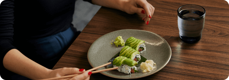 Person eating avocado sushi rolls with chopsticks at a wooden table, accompanied by wasabi, pickled ginger, and a glass of water.
