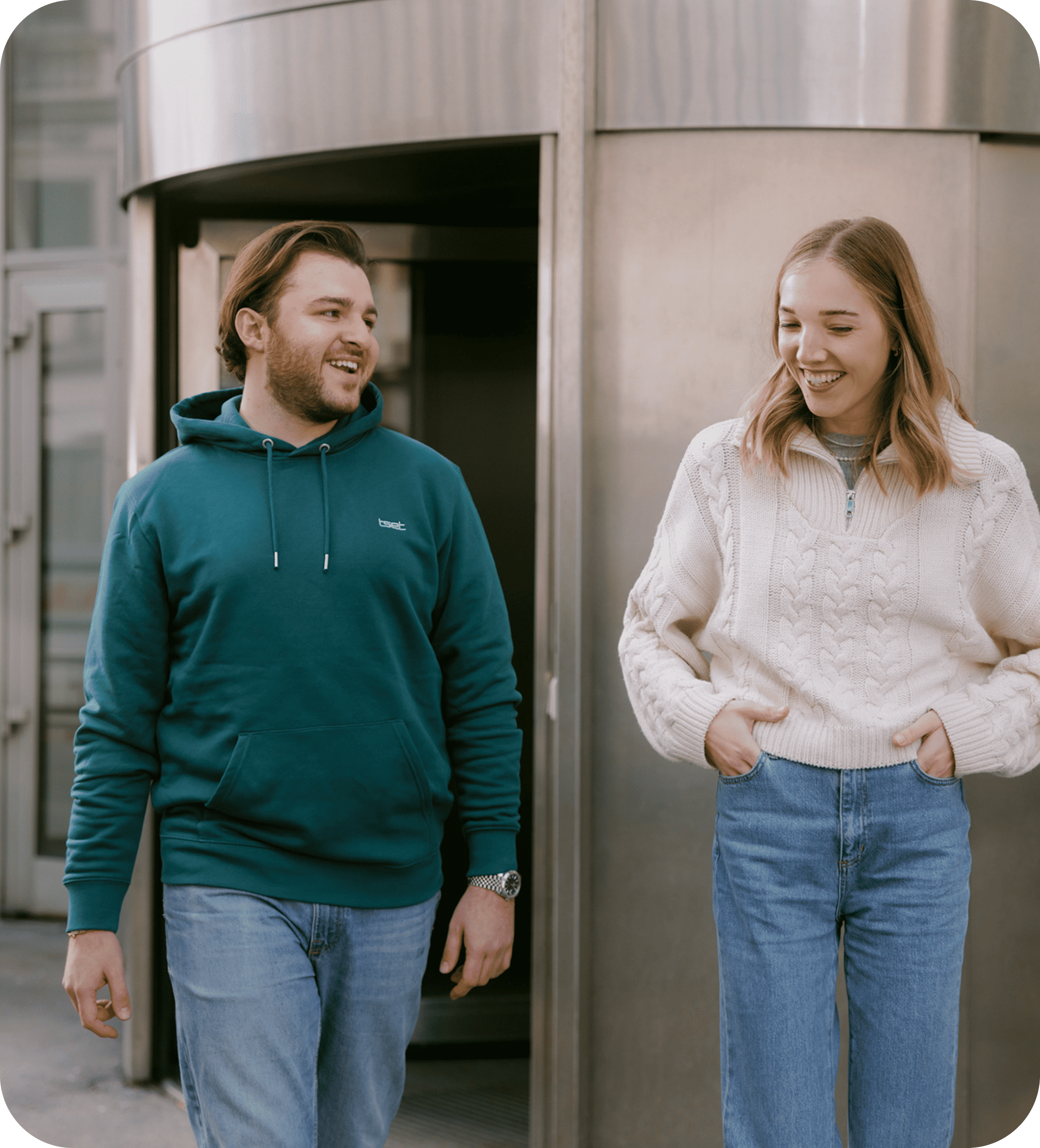 Smiling man and woman walking and chatting in front of a modern office building, dressed casually.