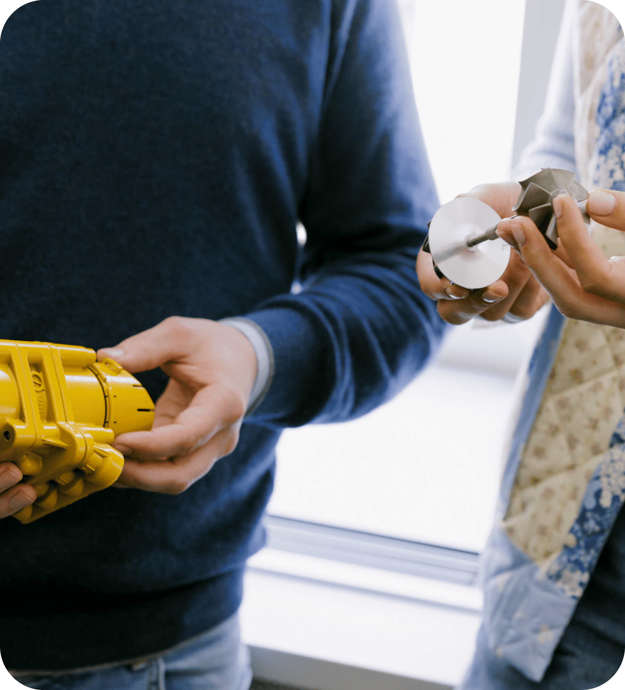Two people examining industrial components, one holding a yellow mechanical part and the other a metal turbine-like piece.