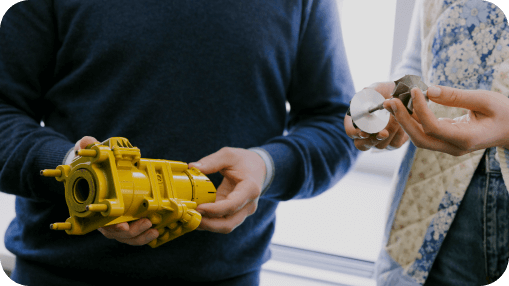 Two people examining industrial components, one holding a yellow mechanical part and the other a metal turbine-like piece. Two people examining industrial components, one holding a yellow mechanical part and the other a metal turbine-like piece.