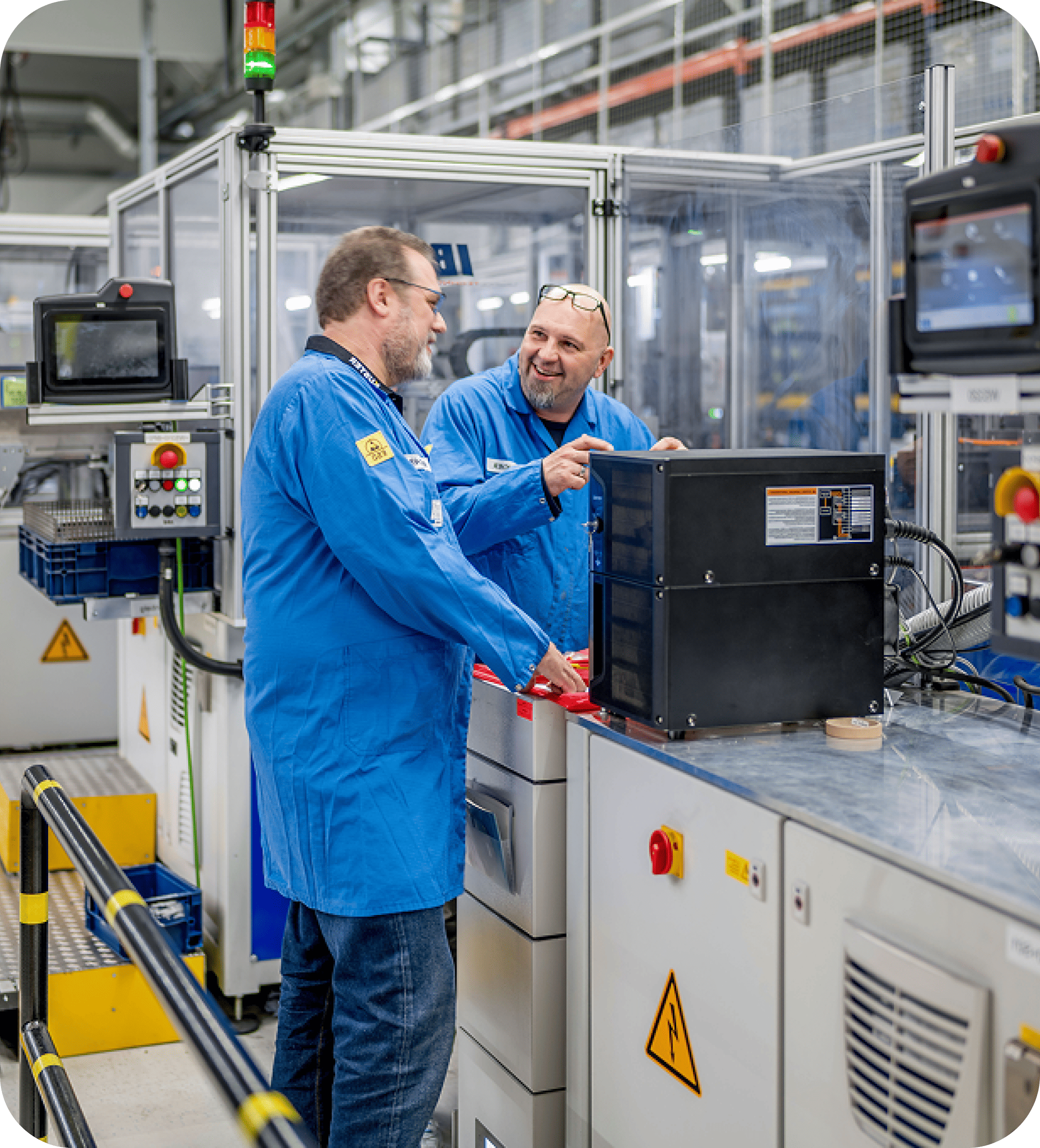 Two engineers in blue lab coats discussing a component at an automated workstation in a modern manufacturing facility.