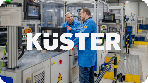Küster logo in front of the two engineers in blue lab coats discussing a component at an automated workstation in a modern manufacturing facility. Küster logo in front of the two engineers in blue lab coats discussing a component at an automated workstation in a modern manufacturing facility.