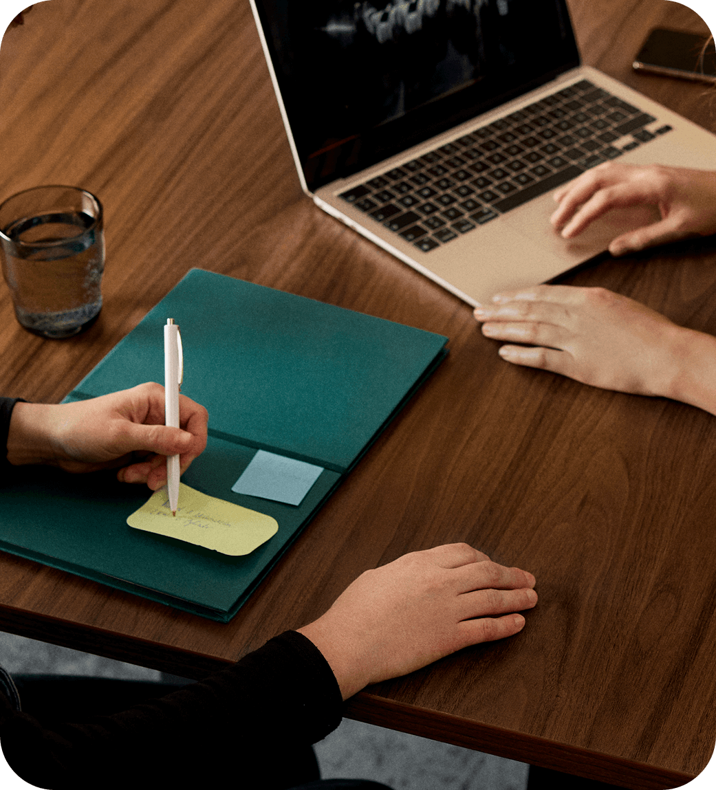 Two people in a meeting at a wooden table with a laptop, notes, and a glass of water, one writing on a notepad.