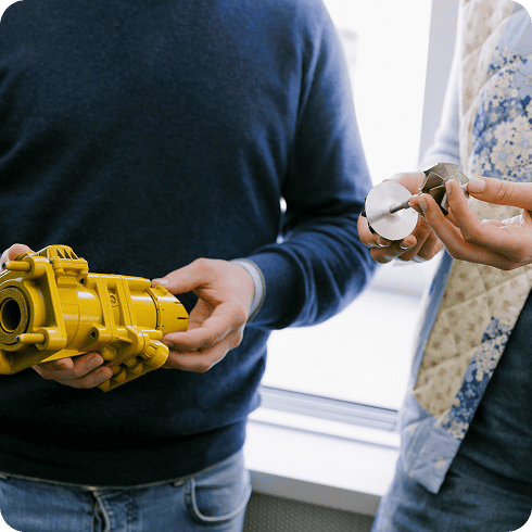Two people examining industrial components, one holding a yellow mechanical part and the other a metal turbine-like piece. Two people examining industrial components, one holding a yellow mechanical part and the other a metal turbine-like piece.