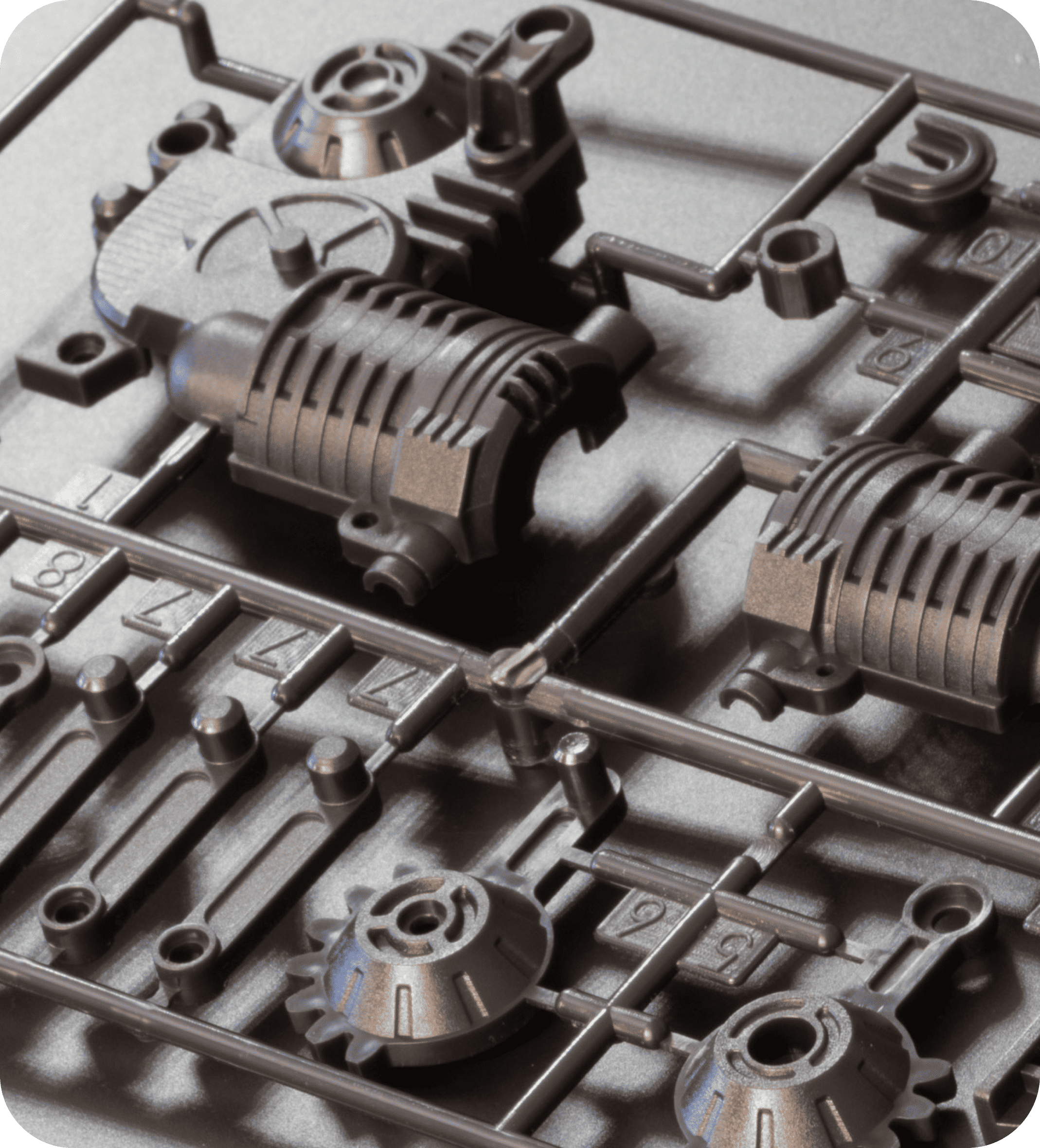 Close-up of plastic model kit components on a sprue, featuring mechanical parts with detailed textures and geometric shapes, in a metallic finish.