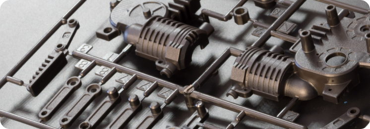Close-up of plastic model kit components on a sprue, featuring mechanical parts with detailed textures and geometric shapes, in a metallic finish.